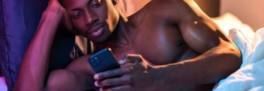 Muscular man lying on a bed and using a smartphone in warm pink and blue light