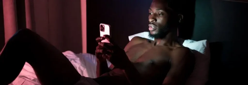 Shirtless man sitting in bed with a laptop in a dimly lit bedroom