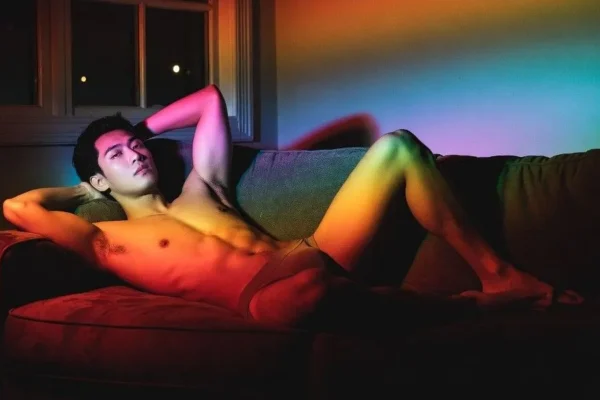 Asian man relaxing on a couch in underwear under colorful rainbow lighting