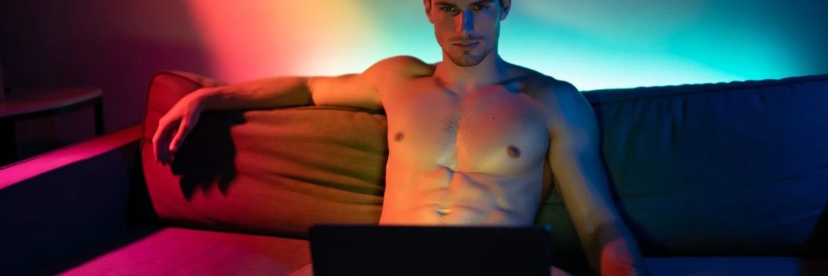 Shirtless man using a laptop on a couch in colorful ambient light