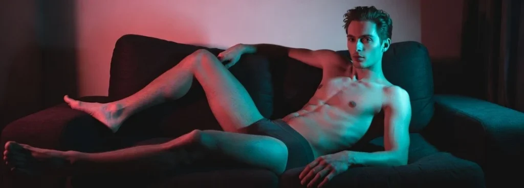 Slim man posing on a couch under dramatic pink and blue lighting